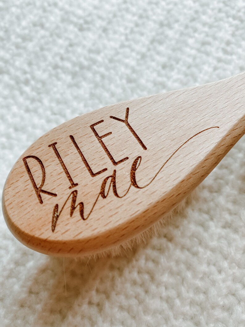 May include: A wooden hair brush with a natural bristle brush head. The handle is engraved with the name "Riley Mae" in a cursive font.
