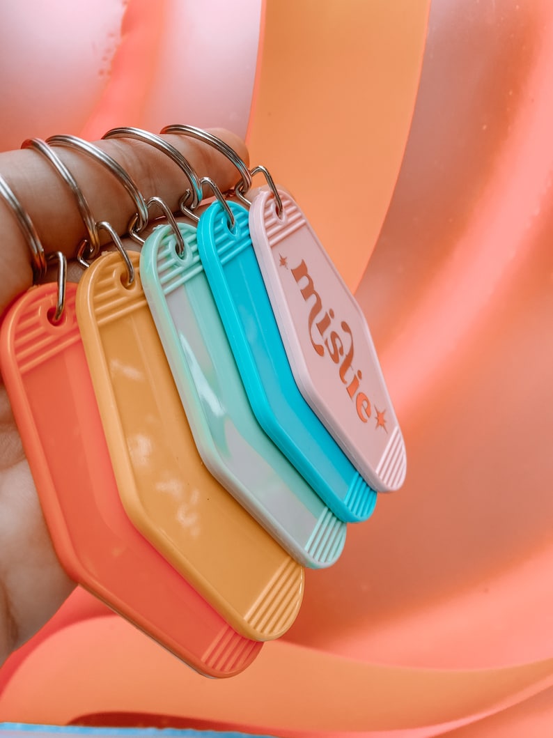 K&ouml;nnte beinhalten: Ein Satz von f&uuml;nf bunten Schl&uuml;sselanh&auml;ngern in Form von Motelzimmerschl&uuml;sseln. Die Schl&uuml;sselanh&auml;nger sind in Orange-, Gelb-, Hellblau-, Rosa- und einem marmorierten Hellblau- und Wei&szlig;ton gehalten. Der rosa Schl&uuml;sselanh&auml;nger tr&auml;gt den Schriftzug "mistie" in goldener Schrift.