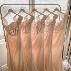 May include: Five peach-colored bridesmaid dresses hanging on a gold rack. The dresses are sleeveless and have a sweetheart neckline. The dresses are all the same style and are made of a soft, flowing fabric.