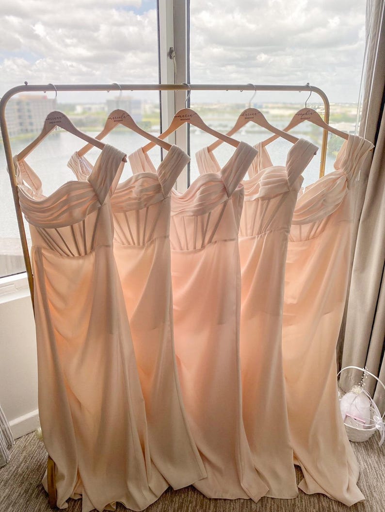 May include: Five peach-colored bridesmaid dresses hanging on a gold rack. The dresses are sleeveless and have a sweetheart neckline. The dresses are all the same style and are made of a soft, flowing fabric.