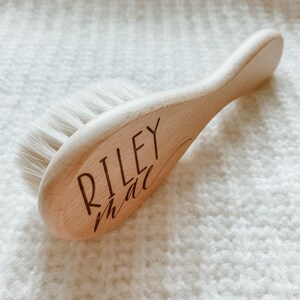 May include: A wooden baby hair brush with soft white bristles. The brush is engraved with the name "Riley" and the word "little" in cursive.