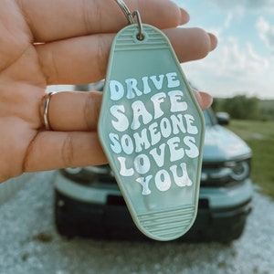 May include: A light green motel-style keychain with the text "DRIVE SAFE SOMEONE LOVES YOU" in white. The keychain is held up in front of a car.