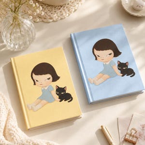 May include: Two hardcover journals, one with a yellow cover and one with a light blue cover, each featuring a graphic illustration of a young person with dark hair in a light blue dress sitting next to a small black cat on the front cover.