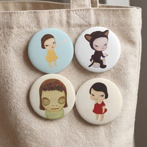 May include: Four round pinback buttons with illustrations of cartoon figures. The buttons feature a girl in a yellow dress, a figure in a cat costume, a girl with large eyes, and a girl in a red dress. The buttons are attached to a beige tote bag.