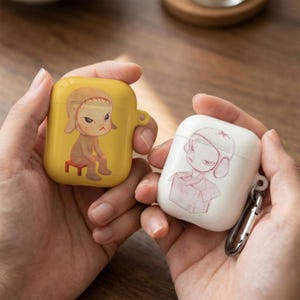 May include: Two AirPod cases, one yellow and one white, each featuring a cartoon illustration. The yellow case has a seated figure with a stern expression. The white case shows a figure inside a box. Both cases have a smooth, glossy finish.