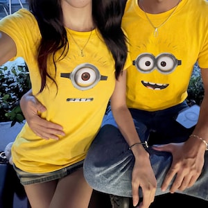 Może przedstawiać: Dwie żółte koszulki z motywem postaci Minionka. Koszulki mają okrągły dekolt i krótkie rękawy. Twarz Minionka jest nadrukowana z przodu, z jednym lub dwoma dużymi oczami i uśmiechniętymi ustami. Koszulki są noszone z dżinsem.