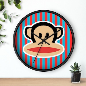 May include: A round wall clock featuring a cartoon monkey face with a red mouth and black ears. The clock face has red and blue vertical stripes. The clock has a black frame and black clock hands.