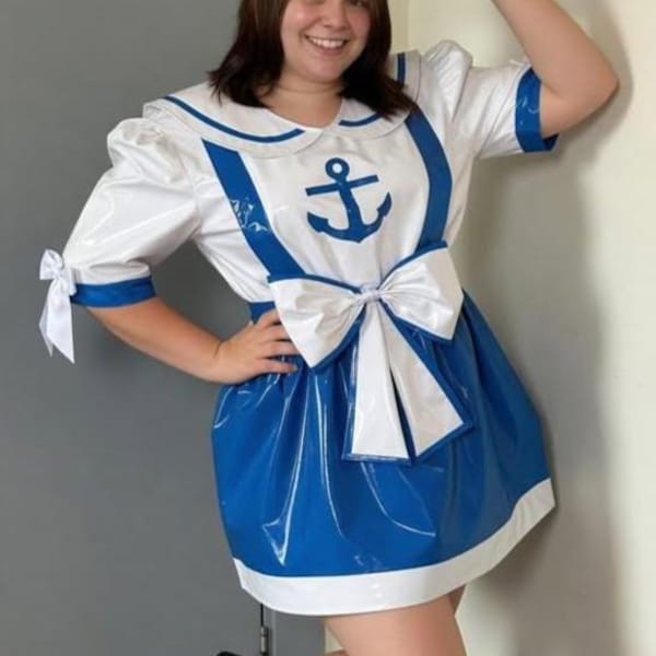 May include: A white and blue sailor costume with an anchor design. The outfit includes a sailor hat, a collared top with an anchor, and a blue skirt. The costume has a large white bow and blue accents. The person is wearing black shoes.