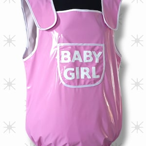 May include: A pink, one-piece garment with white trim and the words "BABY GIRL" printed on the front. The garment has shoulder straps and a pocket. The material appears to be shiny and waterproof.