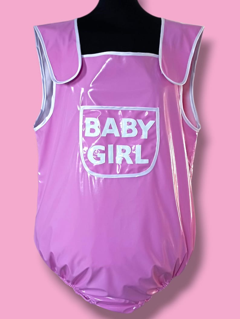 May include: A pink, one-piece garment with white trim and the words "BABY GIRL" printed on the front. The garment has shoulder straps and a rounded bottom, and is made of a shiny material.