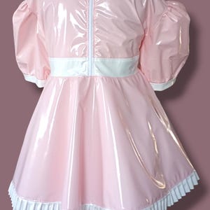 May include: A pink dress with a glossy finish. It has short, puffed sleeves, a white collar, and a white belt. The hemline is trimmed with a white, ruffled edge. A white zip runs down the back.