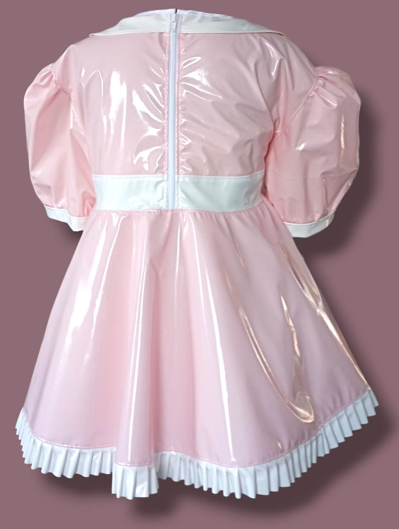 May include: A pink dress with a glossy finish. It has short, puffed sleeves, a white collar, and a white belt. The hemline is trimmed with a white, ruffled edge. A white zip runs down the back.