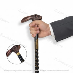 Puede incluir: Un bastón marrón con un mango con forma de cabeza de conejo. El mango es de color bronce y el eje tiene un diseño retorcido. El bastón lo sostiene una persona que lleva un traje.