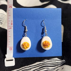 May include: A pair of dangle earrings shaped like halved hard-boiled eggs with white oval whites and textured yellow yolks. The earrings are displayed on a blue card next to a white measuring tape showing a length of approximately 1.25 inches.