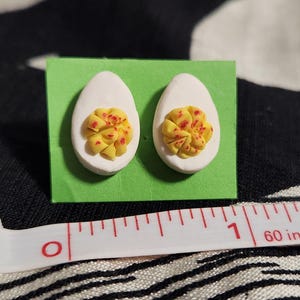 May include: A pair of stud earrings shaped like halved hard-boiled eggs with white egg whites and textured yellow yolks featuring red speckles. The earrings are mounted on a green card next to a white measuring tape showing a scale in inches.
