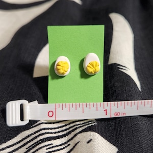 May include: A pair of stud earrings shaped like halved hard-boiled eggs with white whites and textured yellow yolks, mounted on a green card. A white measuring tape is positioned in front, showing the earrings are approximately 0.5 inches wide.
