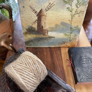 Puede incluir: Un azulejo vintage con una imagen pintada a mano de un molino de viento en un paisaje. El azulejo tiene un esmalte craquelado y un aspecto rústico.