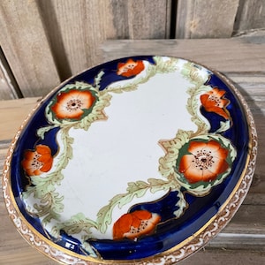 Puede incluir: Un plato ovalado de cerámica con un diseño floral azul y blanco. El plato tiene un borde dorado y presenta flores naranjas, verdes y blancas.