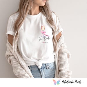 May include: White t-shirt featuring a watercolor illustration of a champagne flute with a pink bow. The words "Team Bride" are written in black script, with a date below. A light-colored cardigan is draped over the shoulders.