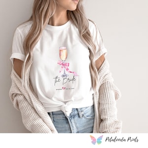 May include: White t-shirt featuring a watercolor illustration of a champagne flute with a pink bow. The text "The Bride" is below the image, along with a date and location. A casual, everyday tee.