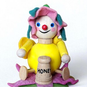 May include: A whimsical wooden doll with a yellow body, arms, and legs. The doll has a wooden head with blue eyes, a red nose, and a smiling mouth. It wears a pink and green flower-shaped hat and holds a wooden honey pot. The word "HONEY" is printed on the pot.