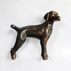 May include: A bronze-colored dog figurine in a standing pose. The sculpture has a glossy finish, with details like the ears, tail, and legs clearly defined. The dog's eyes appear to be a reddish-brown color.