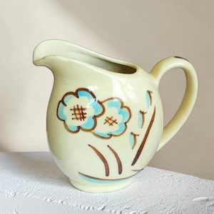 May include: A vintage-style, cream-colored ceramic pitcher with a curved handle and spout. The pitcher is decorated with two blue floral designs with brown crosshatch details and brown stems. The pitcher is sitting on a white surface.