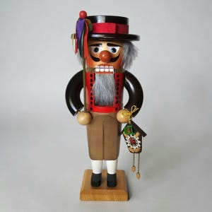 May include: A wooden nutcracker figure wearing a black top hat with a red band and colorful ribbons. The figure has a red jacket, tan trousers, and a gray beard. It holds a miniature cuckoo clock in one hand and stands on a square wooden base.
