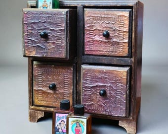 May include: A dark wooden apothecary chest with four drawers, each featuring a textured, embossed metal front with a central black knob. A green bottle sits on top, and two amber glass bottles with vintage-style labels sit in front of the chest.