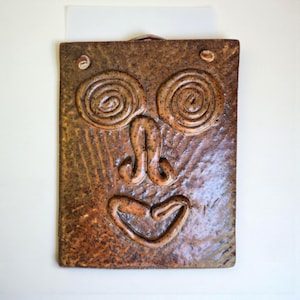 May include: A rectangular, brown ceramic wall hanging with a stylized face design. The face features spiral eyes, a curved nose, and a smiling mouth. The ceramic has a textured surface and is mounted on a white background.