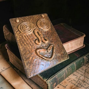 May include: A rectangular brown ceramic plaque featuring a raised, abstract face design with two spiral eyes, a curved nose, and a wide, smiling mouth. The surface has a mottled, textured finish with subtle variations in tone and depth.