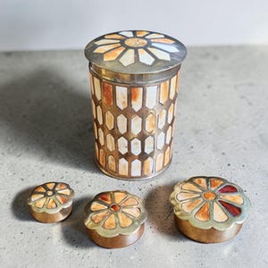 May include: A collection of four decorative boxes with lids. The largest box is cylindrical, featuring a geometric pattern of white and orange segments. The lids have a floral design with orange, white, and red accents. The boxes have a brass-coloured metal trim.