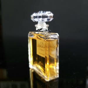 May include: A small, rectangular perfume bottle with a clear, faceted stopper. The bottle contains a golden-colored liquid and has a label with text. The bottle is on a reflective surface.