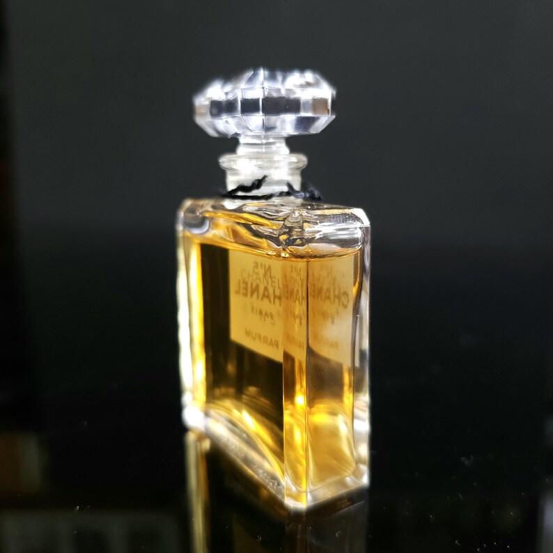 May include: A small, rectangular perfume bottle with a clear, faceted stopper. The bottle contains a golden-colored liquid and has a label with text. The bottle is on a reflective surface.