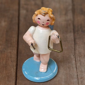 May include: A vintage figurine of an angel playing a triangle. The angel has blonde hair, rosy cheeks, and is wearing a white dress. It stands on a light blue circular base. The triangle is gold-colored.