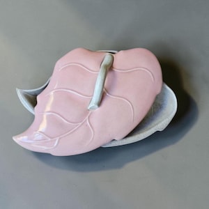May include: A ceramic lidded dish shaped like a leaf. The lid is a smooth, glossy pastel pink with embossed vein details and a curved, light gray handle. The base is a textured, speckled gray leaf shape that extends slightly beyond the pink lid.