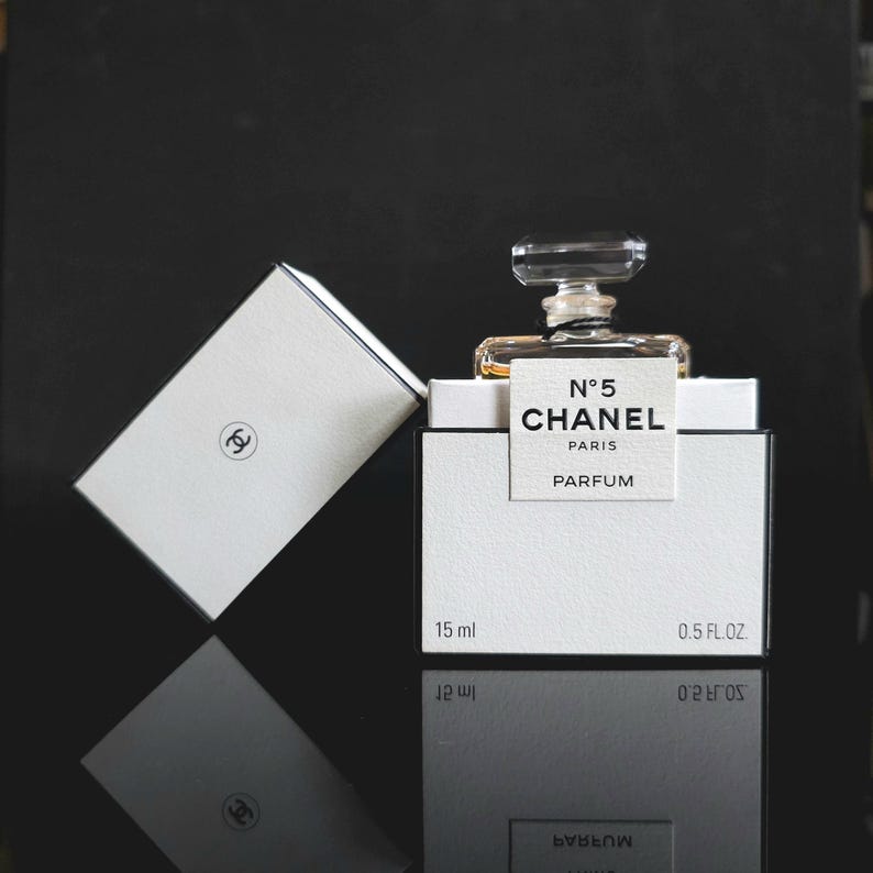 May include: A small bottle of Chanel No. 5 perfume, displayed with its white box. The box and bottle are on a reflective black surface. The box has the Chanel logo and the text "N&deg;5 CHANEL PARIS PARFUM". The bottle contains 15 ml or 0.5 FL.OZ.