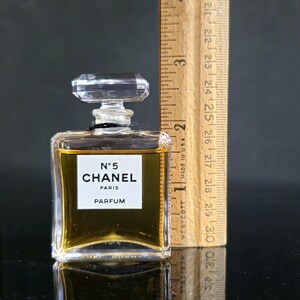 May include: A small, rectangular perfume bottle of Chanel No. 5 Parfum. The clear glass bottle has a faceted stopper and a white label with black text. The liquid inside is amber-colored. A wooden ruler is next to the bottle, measuring approximately 2 inches.