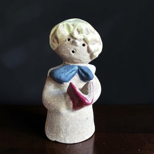 May include: A ceramic figurine of a choirboy, holding a red book. The figurine has a light beige body, a blue bow tie, and a light yellow hairstyle. The background is dark.