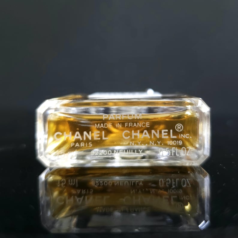 May include: A small, rectangular perfume bottle with a clear glass body and a golden liquid inside. The bottle has the CHANEL logo and text that reads "PARFUM MADE IN FRANCE". The bottle is reflected on a dark surface.