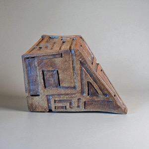 May include: A geometric, abstract ceramic sculpture with a brown and blue color scheme. The piece has a rectangular base and a triangular top, with carved lines and shapes. The sculpture is a unique piece of art.