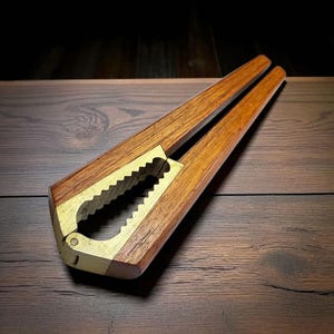 May include: A wooden nutcracker with a brass-colored metal jaw. The nutcracker has a rectangular body with angled sides and a hinged design. The wood has a warm, brown tone, and the metal jaw has a serrated edge.