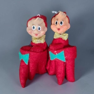 Könnte beinhalten: Zwei Vintage-Wichtelfiguren mit geformten Kunststoffgesichtern, bemalten Merkmalen und blondem Haar. Jeder Wichtel trägt eine rote Filzmütze und einen roten Filzkörper, eine goldene Glitzerfliege am Hals und eine türkisfarbene Filzschleife vorne.