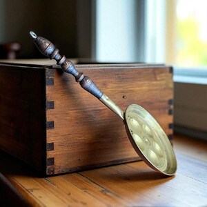 May include: A vintage brass egg poacher with a wooden handle. The handle is dark brown with decorative rings. The poacher has a circular cooking surface with six indentations. The brass has a warm, golden hue, and the wood has a rich, dark tone.