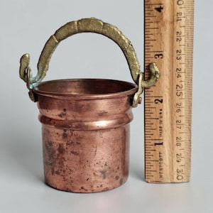 May include: A small copper bucket with a textured brass handle, positioned next to a wooden ruler showing a height of approximately 2.5 inches. The copper surface displays a weathered, mottled patina with visible dark spots and a cylindrical shape.