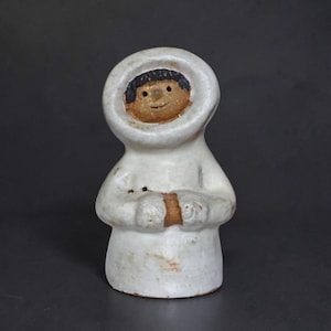May include: A ceramic figurine of a person in a white hooded parka. The face is tan with black hair and a smiling expression. The figure's hands are clasped in front, and the body is a solid white color.