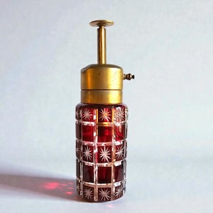 May include: A vintage red glass perfume bottle with a brass top and stem. The bottle has a geometric pattern with starburst designs. The brass components include a small nozzle and a circular top. The bottle is likely from the mid-century era.