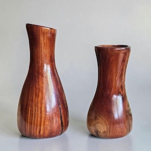 May include: Two polished wooden vases with a smooth, glossy finish and visible natural grain patterns. The taller vase on the left features a slightly angled rim, while the shorter vase on the right has a rounded, symmetrical opening and base.