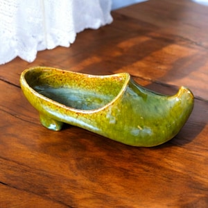 May include: A glazed ceramic shoe-shaped planter in green and yellow hues. The planter, with its curved toe and small heel, is placed on a wooden surface. It's designed for small plants or decorative items, and has a glossy finish.