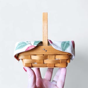 May include: A small, brown wicker basket with a fabric liner featuring a red apple and green leaf pattern. The basket has a wooden handle and is perfect for carrying small items.